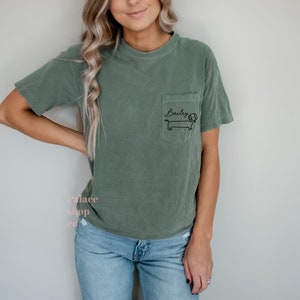 May include: A green t-shirt with a pocket on the left side. The pocket has a black dachshund with the name "Bailey" written above it.