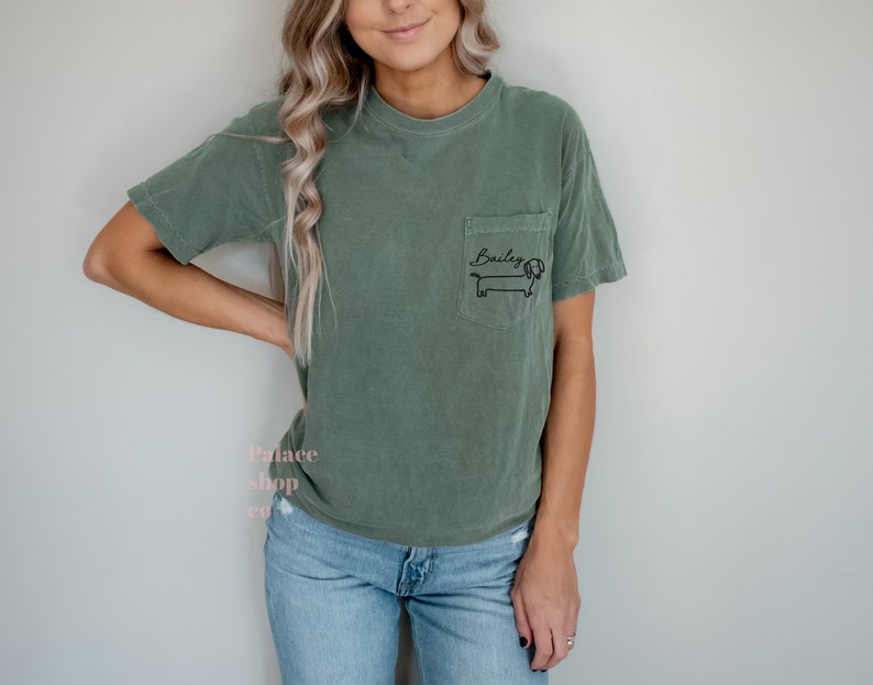 May include: A green t-shirt with a pocket on the left side. The pocket has a black dachshund with the name "Bailey" written above it.