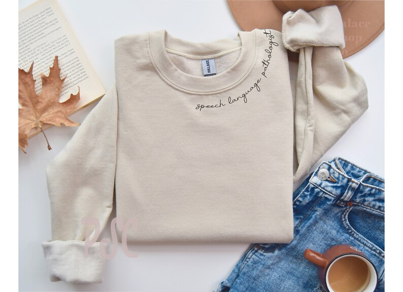 Puede incluir: Una sudadera beige con el texto "speech language pathologist" escrito en cursiva negra en el escote.