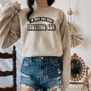 May include: A beige sweatshirt with the text "I'm just out here trusting God" in black lettering.