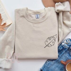 May include: A beige sweatshirt with a simple line drawing of a leaf on the left side of the chest.