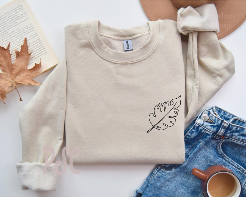 May include: A beige sweatshirt with a simple line drawing of a leaf on the left side of the chest.