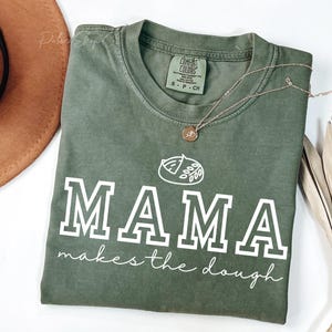 Pode incluir: Uma camiseta verde com o texto "MAMA makes the dough" em branco. A camiseta tem uma pequena ilustração de uma fatia de pizza com um contorno branco.