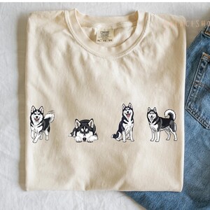 Pu&ograve; includere: T-shirt color crema con quattro cani husky dei cartoni animati in diverse pose. I cani sono neri e bianchi con occhi blu. La maglietta &egrave; mostrata con jeans blu.