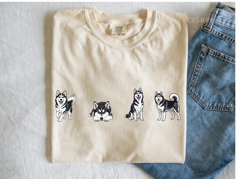 Pu&ograve; includere: T-shirt color crema con quattro cani husky dei cartoni animati in diverse pose. I cani sono neri e bianchi con occhi blu. La maglietta &egrave; mostrata con jeans blu.