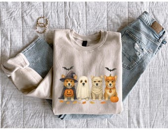 May include: A beige sweatshirt featuring a Halloween-themed design with four corgi dogs dressed in costumes: a witch, a ghost, a mummy, and a pumpkin. The design includes bats and autumn leaves. The sweatshirt is paired with light blue jeans and gold hoop earrings.