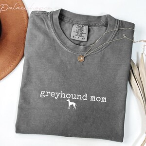 May include: A gray Comfort Colors t-shirt with the words "greyhound mom" and a greyhound silhouette in white. A gold pendant necklace and a brown hat are also in the image.