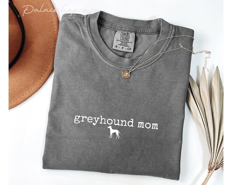 May include: A gray Comfort Colors t-shirt with the words "greyhound mom" and a greyhound silhouette in white. A gold pendant necklace and a brown hat are also in the image.