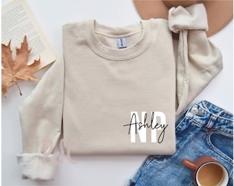 Puede incluir: Una sudadera beige con un diseño en blanco y negro que dice "Ashley NP" en la parte delantera.
