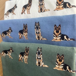 Op de afbeelding: Een stapel gevouwen sweatshirts in wit, blauw en groen, elk met een gedetailleerde illustratie van een Duitse herdershond in verschillende poses. De honden zijn afgebeeld in zwart, bruin en beige.