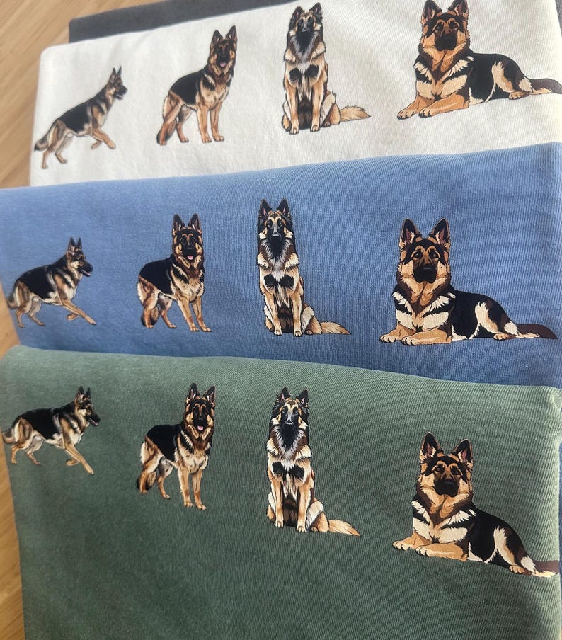 Op de afbeelding: Een stapel gevouwen sweatshirts in wit, blauw en groen, elk met een gedetailleerde illustratie van een Duitse herdershond in verschillende poses. De honden zijn afgebeeld in zwart, bruin en beige.