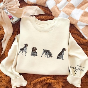 Peut inclure: Sweat-shirt crème avec quatre illustrations de chiens dans différentes poses. Les chiens sont dans des tons de marron, noir et gris. La manche droite porte le mot "custom" en écriture manuscrite.
