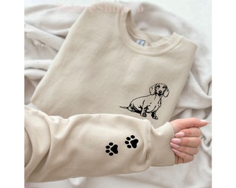 Könnte beinhalten: Ein beiger Pullover mit einer schwarzen Dackel-Silhouette auf der Vorderseite und zwei schwarzen Pfotenabdrücken auf dem linken Ärmel.