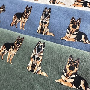 Op de afbeelding: Close-up van drie stoffen stalen in wit, blauw en groen, elk bedrukt met herhaalde illustraties van Duitse herders in verschillende poses. De honden zijn afgebeeld in zwart, bruin en bruin.