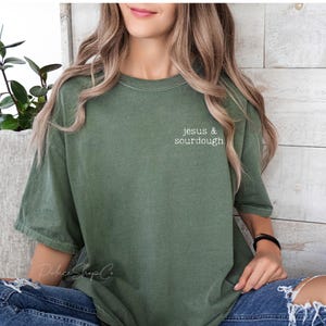 May include: A woman wearing a green t-shirt with the text "jesus & sourdough" in white lettering. She is wearing blue jeans and has long brown hair.