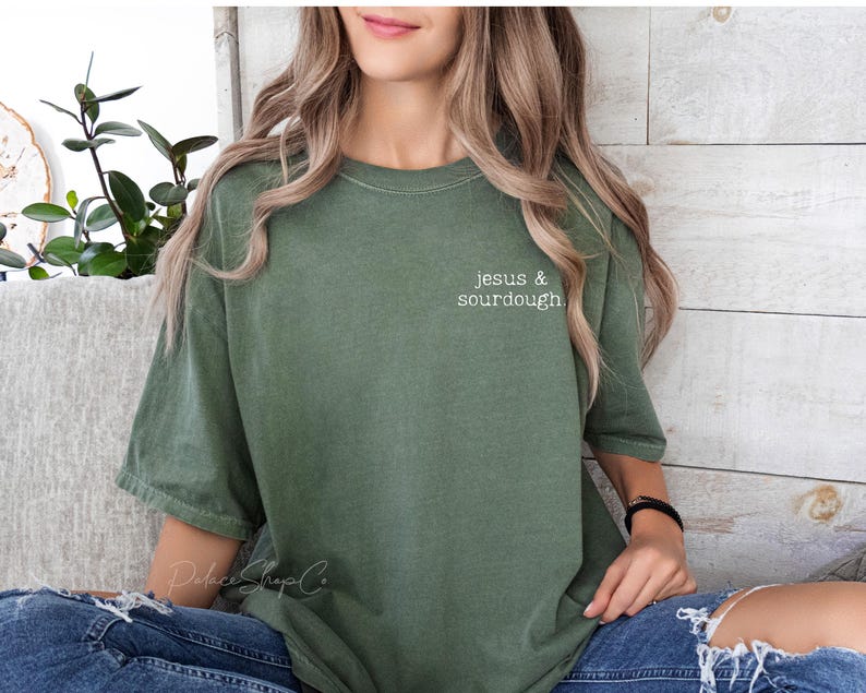 May include: A woman wearing a green t-shirt with the text "jesus & sourdough" in white lettering. She is wearing blue jeans and has long brown hair.