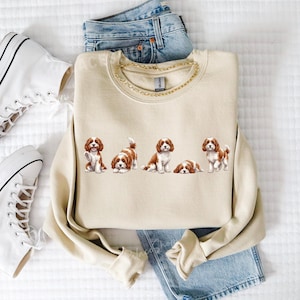 Op de afbeelding: Een beige sweatshirt met illustraties van Cavalier King Charles Spaniels in verschillende poses. Het sweatshirt wordt gecombineerd met een blauwe jeans en witte high-top sneakers. Een gouden kettingketting accentueert de halslijn.
