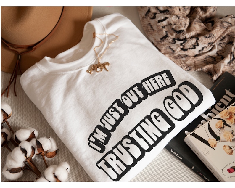 May include: White sweatshirt with the text "I'm just out here trusting God" in a bold, black, outlined font. The sweatshirt is laid out on a surface with a brown hat, a patterned scarf, and a book.
