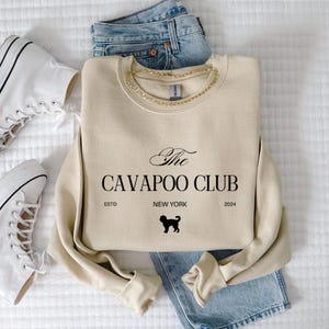 May include: Cream-colored sweatshirt with "The Cavapoo Club" text, a dog silhouette, and "New York 2024" printed on it. The sweatshirt is paired with blue jeans, white sneakers, and a gold chain.