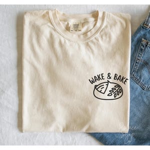 Può includere: Una maglietta color crema con la scritta "Wake & Bake" e un'illustrazione di un dolce con un morso.