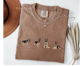 May include: A brown t-shirt featuring four embroidered German Shepherd dog designs in various poses. The shirt has a crew neck and short sleeves. A silver necklace with a pendant is draped across the shirt. The brand tag reads "Comfort Colors".