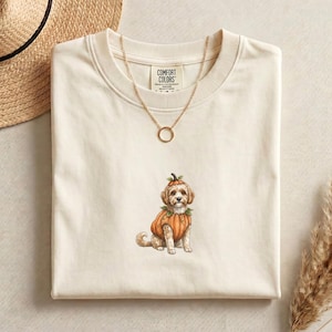 Può includere: Una t-shirt a maniche corte color crema con la grafica di un cane marrone chiaro che indossa un costume da zucca. La maglietta presenta un'etichetta del marchio Comfort Colors sul collo interno ed è abbinata a una sottile collana dorata.