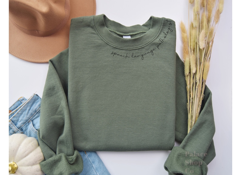 Puede incluir: Una sudadera verde con el texto "speech language pathologist" escrito en cursiva negra en la parte delantera.