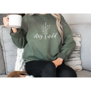 May include: A green sweatshirt with the text "stay wild" and a white floral design. The sweatshirt is being worn by a person sitting on a couch.