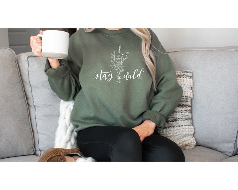 May include: A green sweatshirt with the text "stay wild" and a white floral design. The sweatshirt is being worn by a person sitting on a couch.