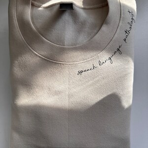 Puede incluir: Una sudadera beige con el texto "speech language pathologist" bordado en la parte delantera.