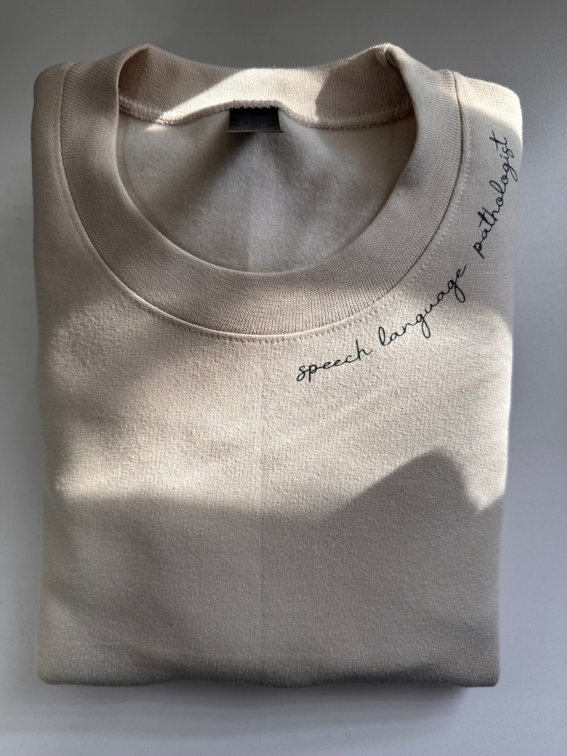 Puede incluir: Una sudadera beige con el texto "speech language pathologist" bordado en la parte delantera.