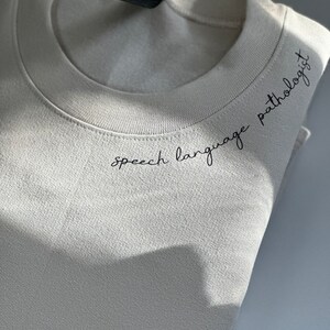 Puede incluir: Una sudadera beige con el texto "speech language pathologist" bordado en la parte delantera.