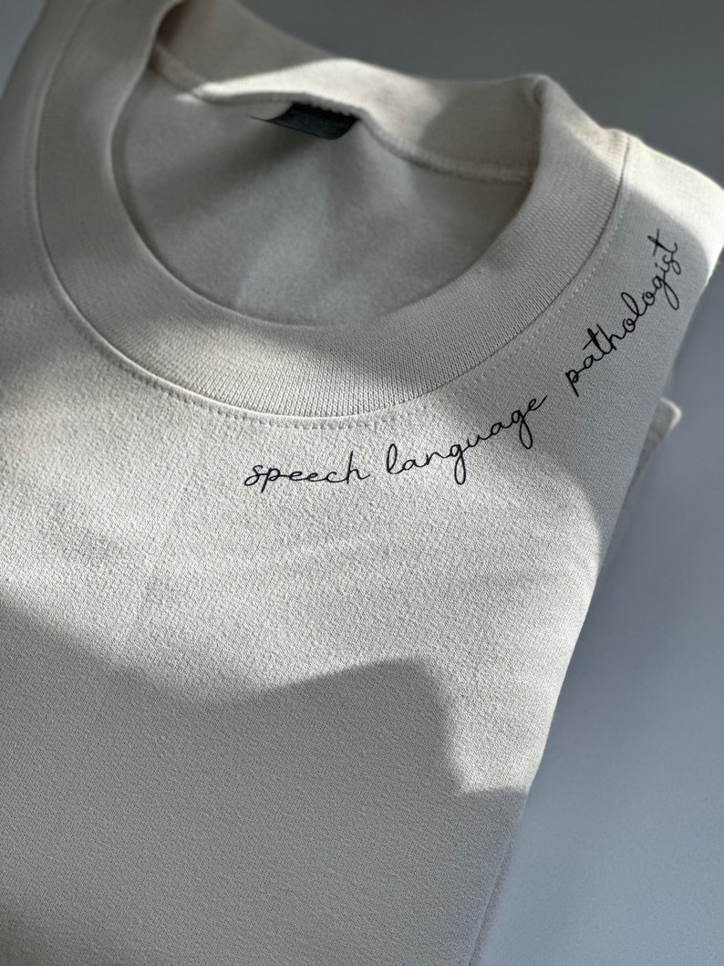 Puede incluir: Una sudadera beige con el texto "speech language pathologist" bordado en la parte delantera.