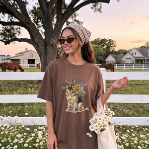 Peut inclure: T-shirt marron avec un motif de berger allemand tenant un bouquet de tournesols. La personne porte des lunettes de soleil et un foulard &agrave; motifs, tenant un sac fourre-tout avec des marguerites. L'arri&egrave;re-plan comprend une cl&ocirc;ture blanche, des chevaux et une grange.