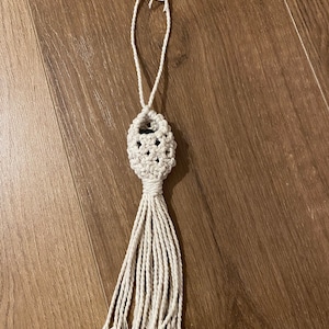May include: A white macrame tassel with a knotted oval shape at the top. The tassel is hanging from a white cord.