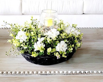 Puede incluir: Un arreglo floral blanco y verde en un tazón negro con un portavelas de vidrio sobre una mesa de café blanca con una alfombra estampada.
