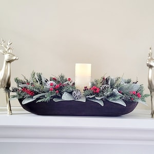 May include: A wooden bowl filled with a Christmas centrepiece featuring greenery, red berries, pine cones, and a white pillar candle. Two gold deer figurines flank the bowl, and two white candle holders with greenery and red berries are on either side of the bowl. The centrepiece is on a white mantel.