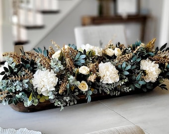 May include: A long, dark brown wooden dough bowl filled with an arrangement of white flowers, dark green eucalyptus leaves, and textured golden-brown dried botanical accents, creating a dense, horizontal centerpiece for a dining table.