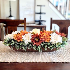 May include: A wooden dough bowl filled with a floral arrangement featuring white and orange flowers, green foliage, and a white candle in the center.