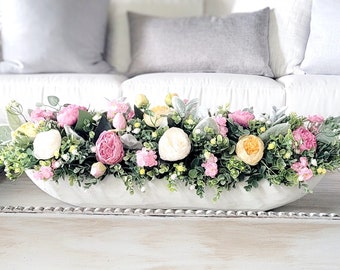 May include: A white ceramic trough planter filled with a floral arrangement of pink, white, and yellow flowers. The planter is sitting on a white coffee table.