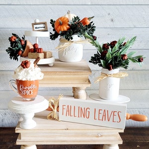 May include: A tiered tray decorated with fall-themed items, including a white mason jar with a brown twine bow, a white mason jar with a brown twine bow filled with greenery and small orange pumpkins, a white tiered tray with a sign that says "Harvest", and a white mug with the text "Pumpkin Spice". The tray is on a wooden table with a sign that says "Falling Leaves".