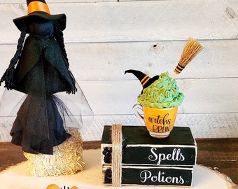 May include: A Halloween-themed decoration featuring a witch figurine, a stack of wooden blocks with the words "Spells" and "Potions", and a beaded garland with orange, white, and black beads. The garland has a black and white striped bow and a brown tassel.