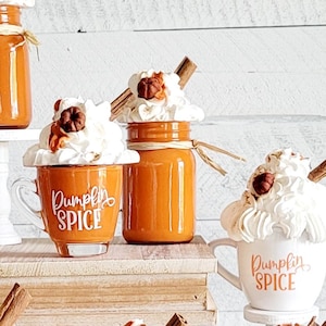 Può includere: Un'esposizione di oggetti decorativi a tema spezie di zucca. L'allestimento comprende tazze e barattoli arancioni sormontati da panna montata, bastoncini di cannella e accenti decorativi. Le tazze hanno il testo "Pumpkin Spice".