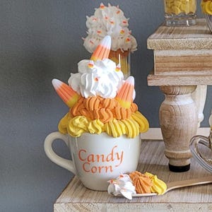 Può includere: Un'esposizione a più livelli di oggetti decorativi a tema caramelle di mais. Include cupcakes, tazze e bicchieri riempiti con strati di giallo, arancione e bianco, sormontati da panna montata e pezzi di caramelle di mais. Le tazze e i bicchieri hanno il testo "Candy Corn".