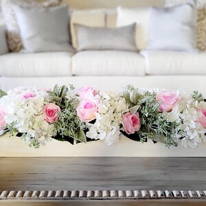 May include: A white rectangular planter filled with a floral arrangement of pink roses and white hydrangeas. The planter is sitting on a wooden table.