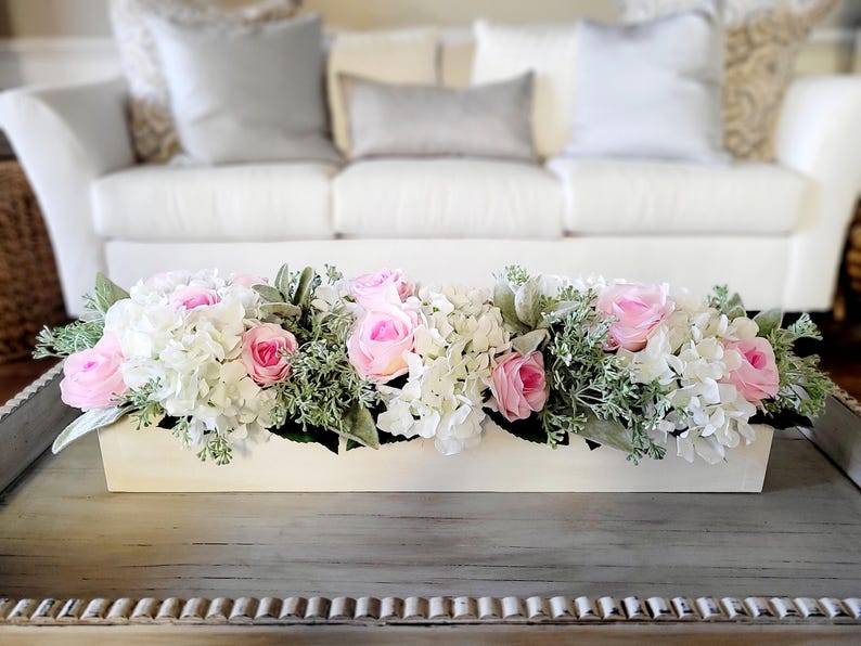 May include: A white rectangular planter filled with a floral arrangement of pink roses and white hydrangeas. The planter is sitting on a wooden table.