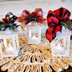 May include: Three white metal lanterns with LED lights, each decorated with a different plaid bow and greenery. The lanterns are sitting on a woven straw mat.