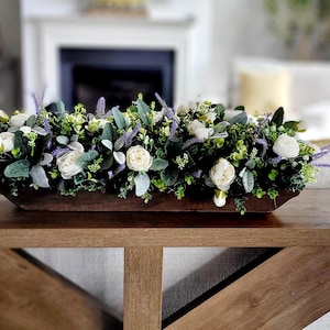 May include: A wooden centerpiece filled with white roses, lavender, and greenery sits on a wooden table. Two gold lamps with white shades flank the centerpiece. The table has a decorative X-shaped support.