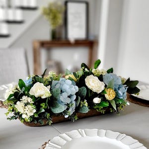 以下が含まれることがあります： ライトブルーの紫陽花、クリームホワイトのバラ、小さな黄色い花、そして様々な緑のユーカリの葉や枝葉で構成された、長方形の木製ボウルに入った人工花のフラワーアレンジメント。