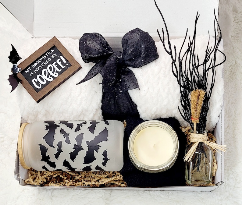 Puede incluir: Una caja de regalo blanca llena de una manta de lana con estampado blanco y negro, una cinta de sat&eacute;n negra, una vela de vidrio, un frasco de vidrio con un patr&oacute;n de murci&eacute;lago negro y una peque&ntilde;a botella de vidrio con una rama marr&oacute;n y un borla marr&oacute;n. La caja tambi&eacute;n incluye un letrero de madera que dice "My broomstick is powered by coffee!"